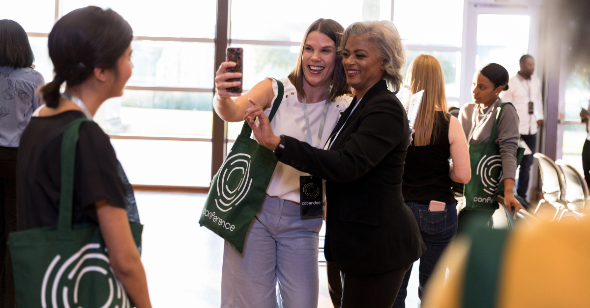 Two women posing for selfie taken by cell phone in the lobby of a work conference, with other people in the background and all wearing conference branded tote bags and clothing.