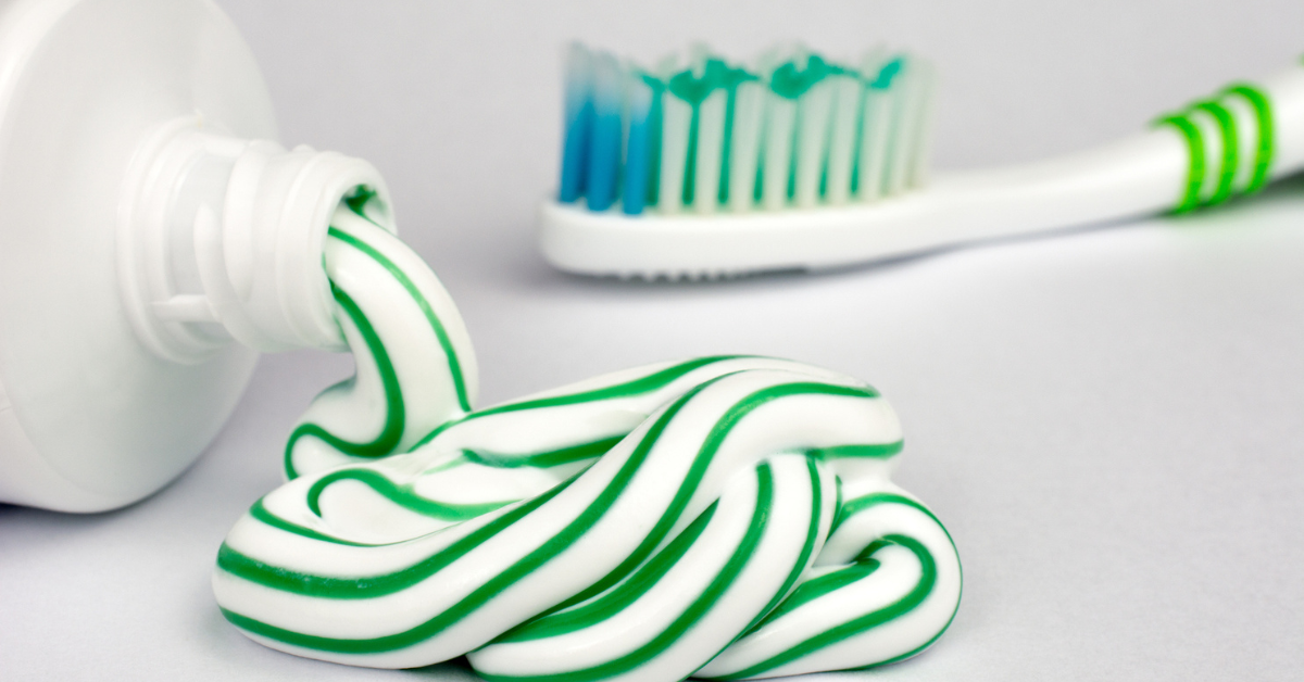 Tube of toothpaste squeezing green and white striped toothpaste out with a green toothbrush in the background.