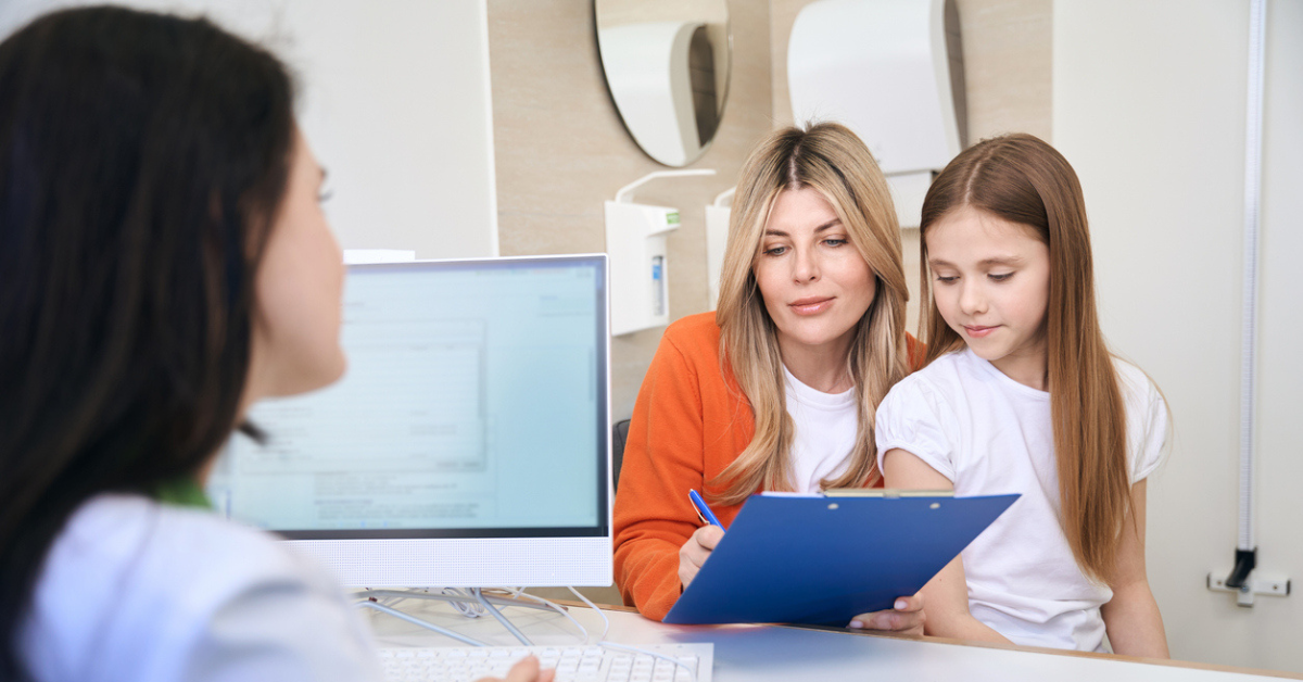 Mother and daughter sitting on other side of desk across from a medical professional. Mother is holding a clipboard and pen, signing paperwork while daughter watches.